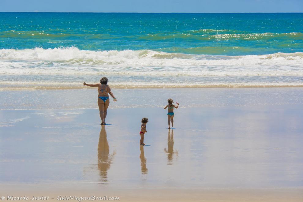 Imagem de uma mãe com suas duas filhas pequenas andando em direção ao mar.